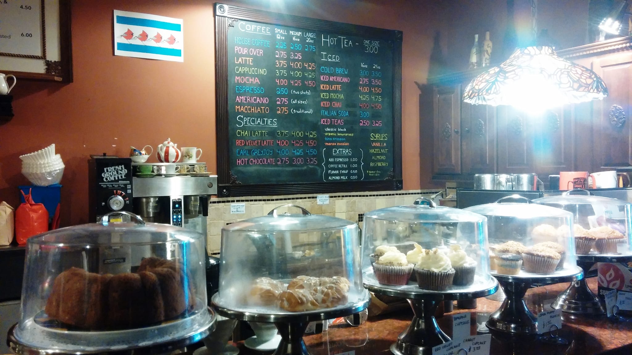 Cake domes of cupcakes and pastries below a colorful chalkboard coffee menu