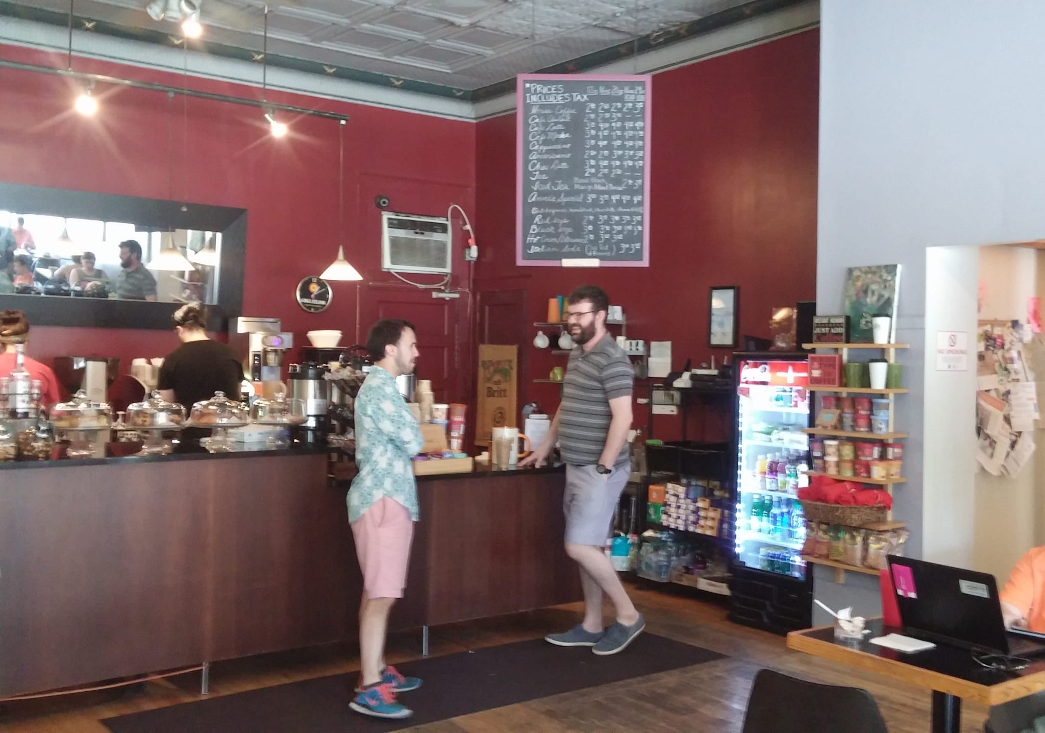 Customers at a wooden counter with pastry domes and chalkboard price list