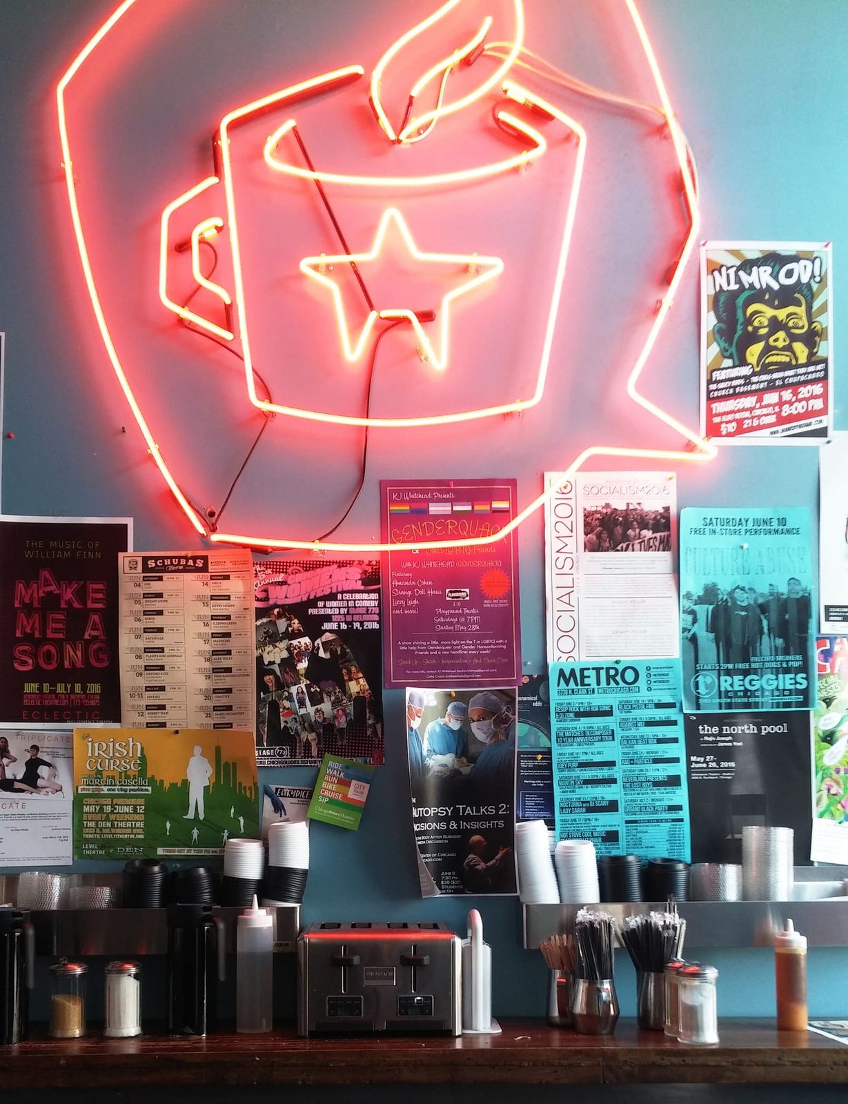 Neon coffee cup sign over a wall of gig posters and condiment counter