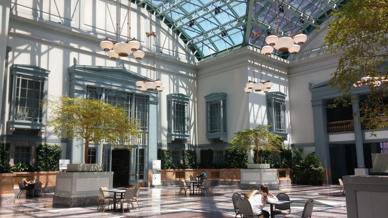 Sunlit glass-roofed atrium with cafe tables, trees and hanging lamps