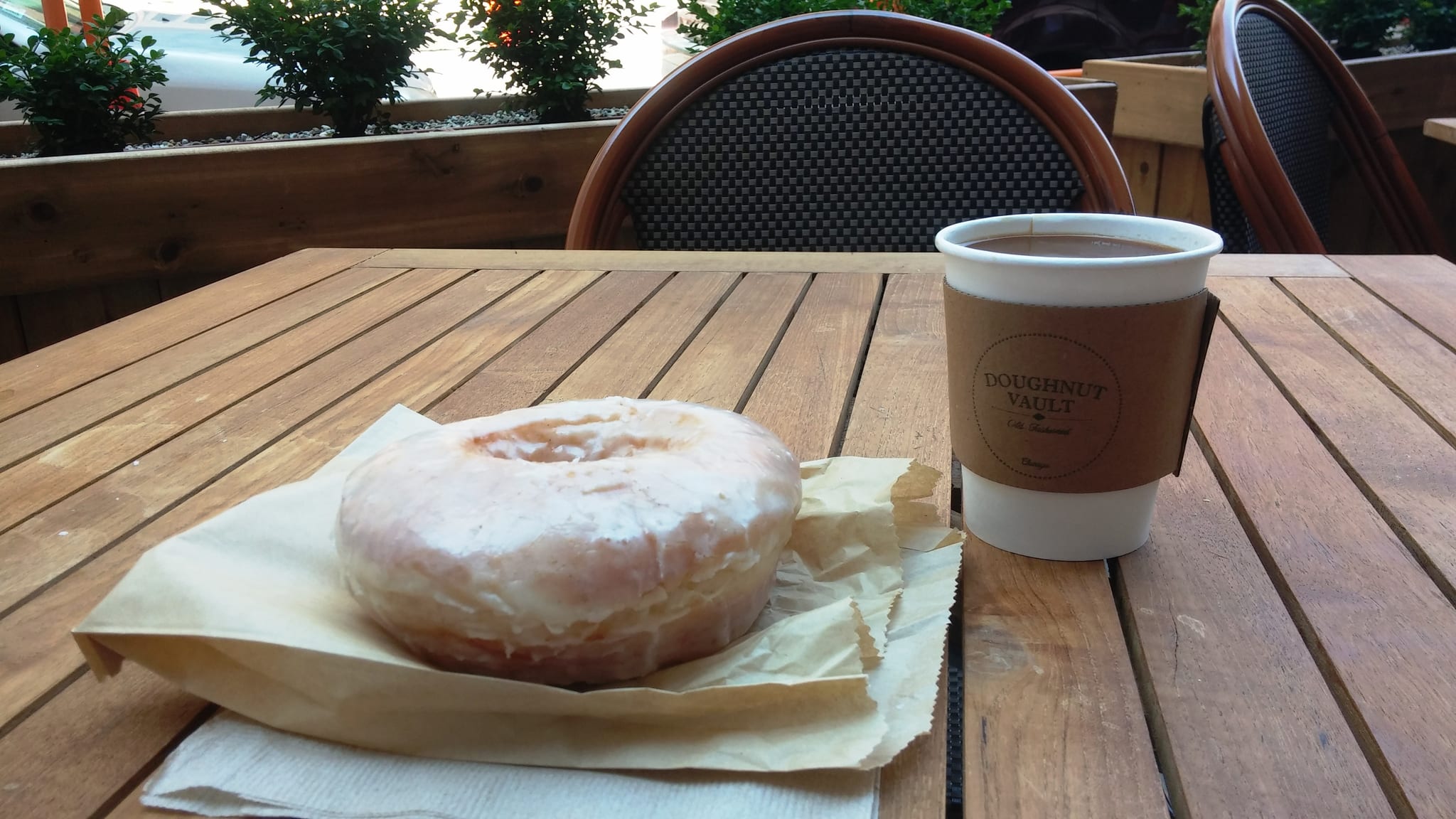 Glazed doughnut on paper beside a paper coffee cup outdoors