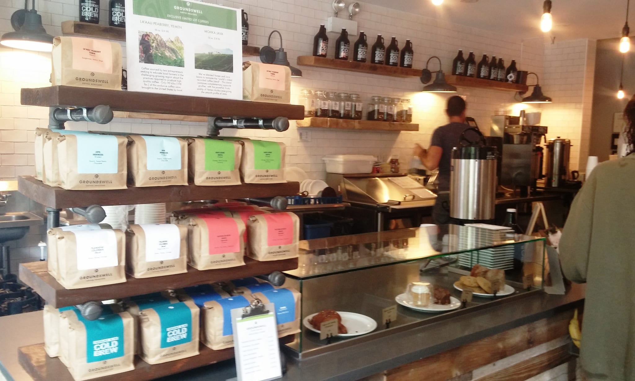 Retail shelves of coffee bags beside pastry case and barista at work