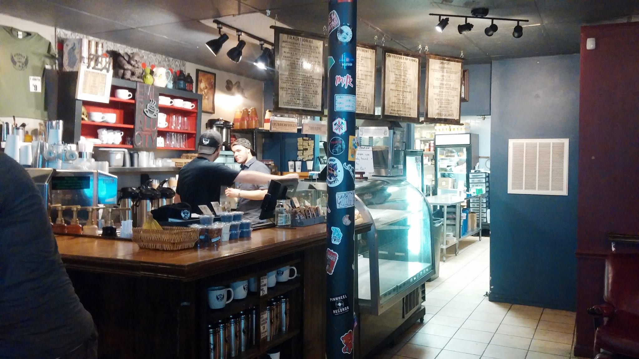 Wooden service counter, pastry case and baristas beneath hanging chalk menu boards