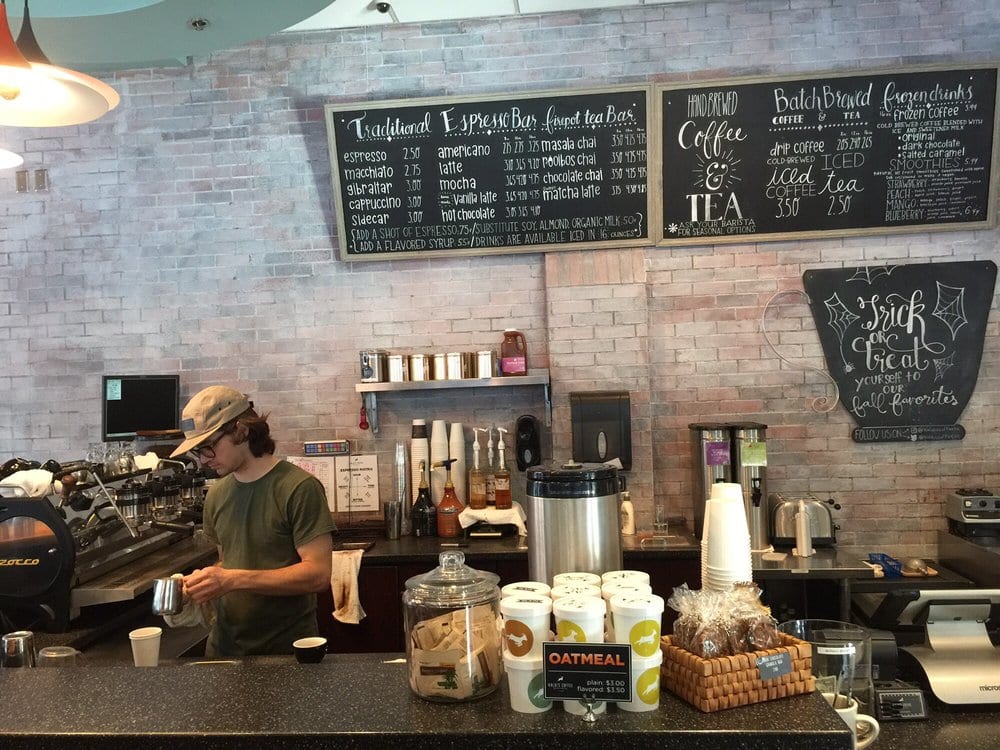Barista steaming milk below handwritten chalkboard menus on whitewashed brick
