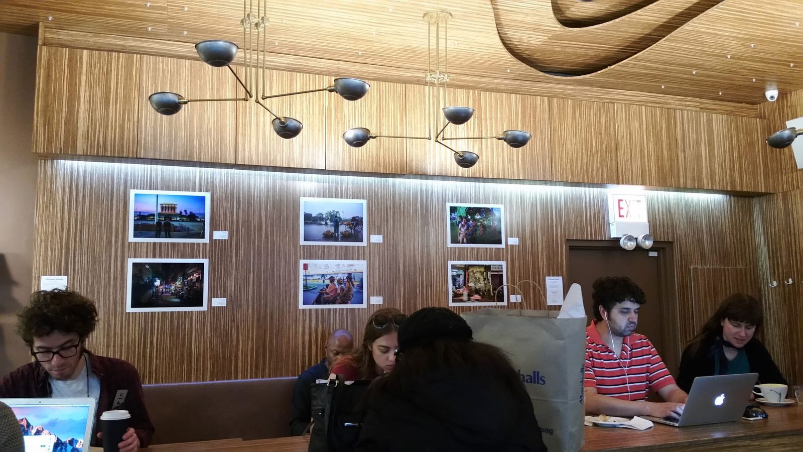 Customers on laptops at a communal table below a photo exhibit on zebrawood walls