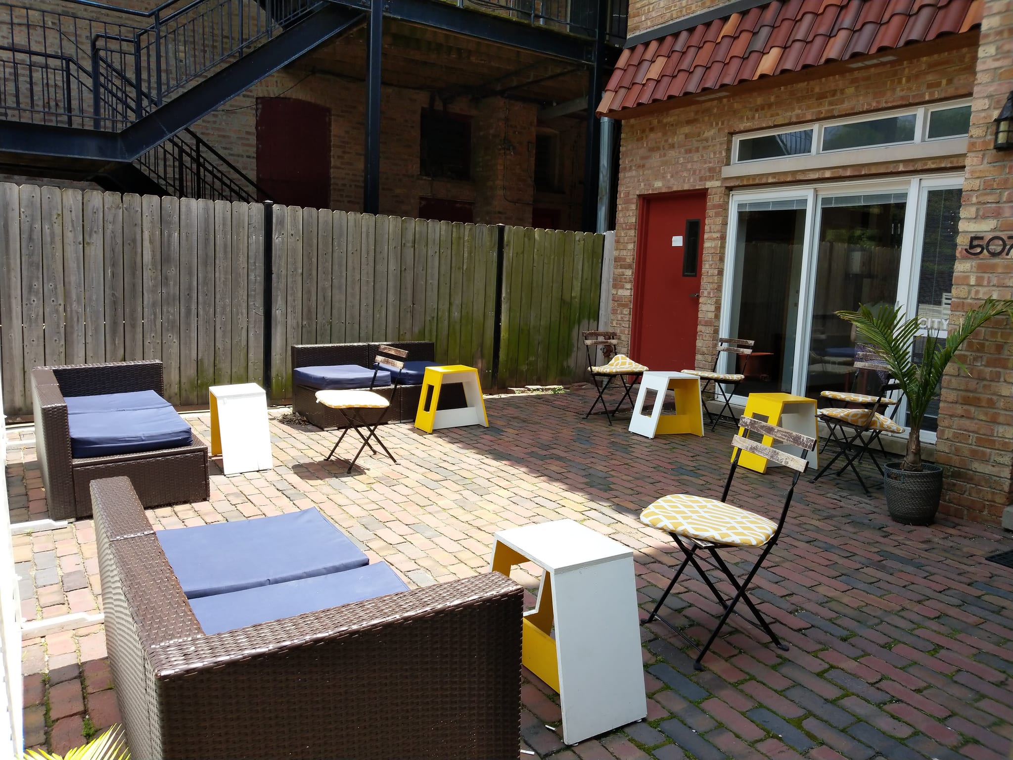 Brick-paved back patio with wicker lounge seating and yellow bistro chairs