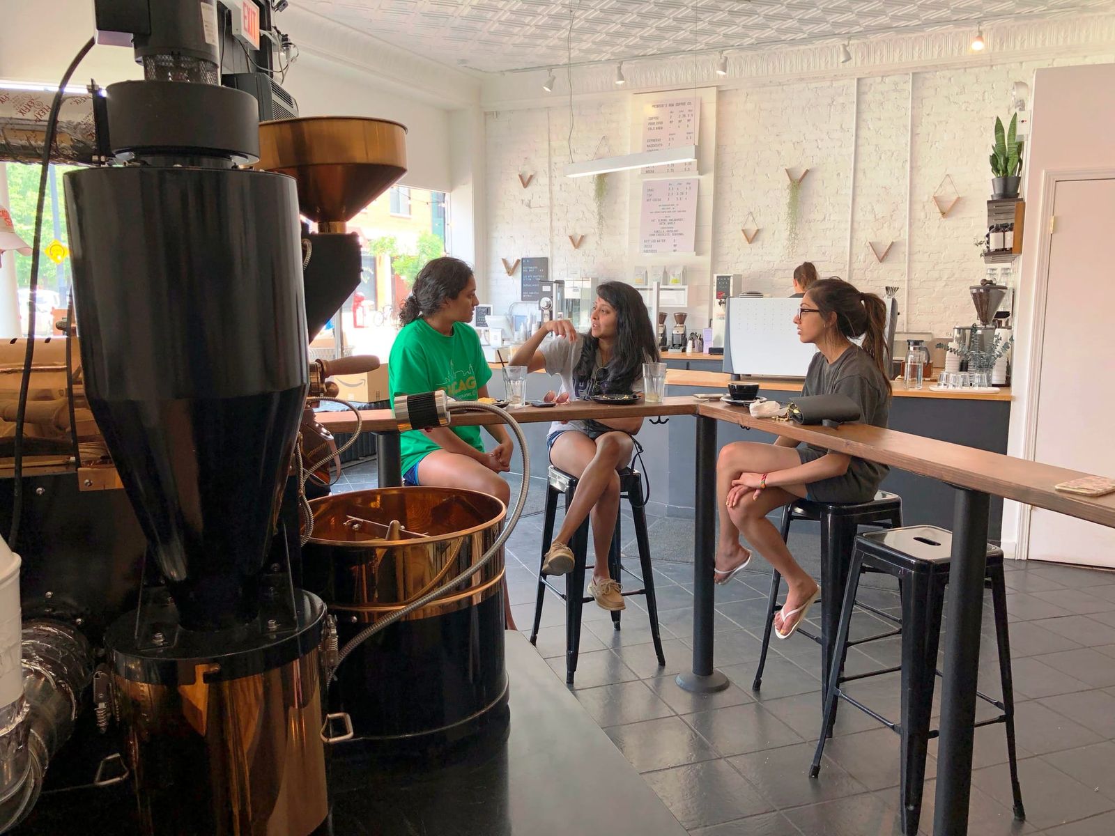 Copper drum coffee roaster beside three women talking at a stool-lined bar