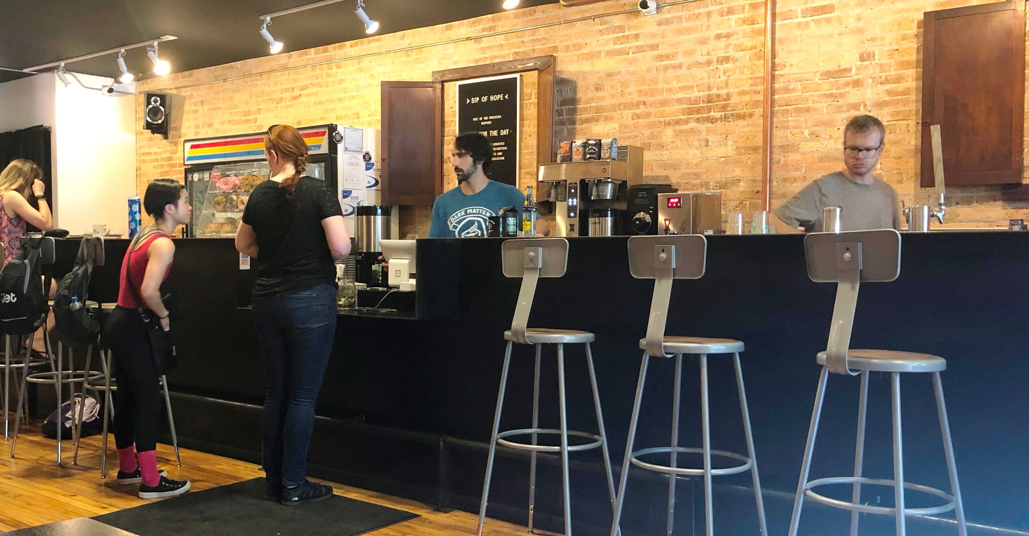 Customers ordering at a dark counter under exposed brick, tall silver stools