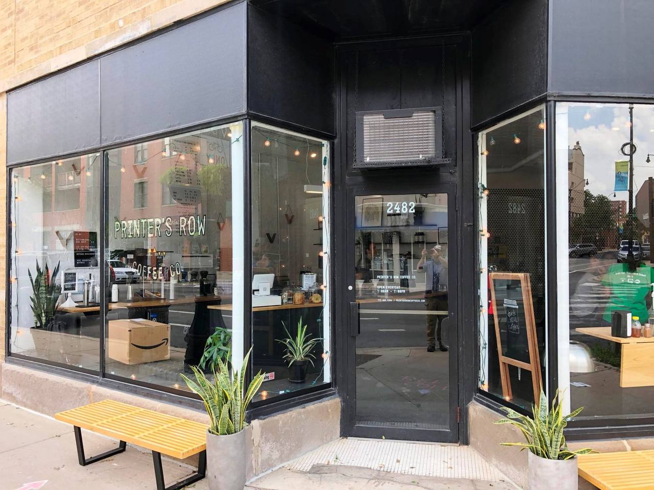 Corner storefront with black trim, painted window signage, bench and potted plants