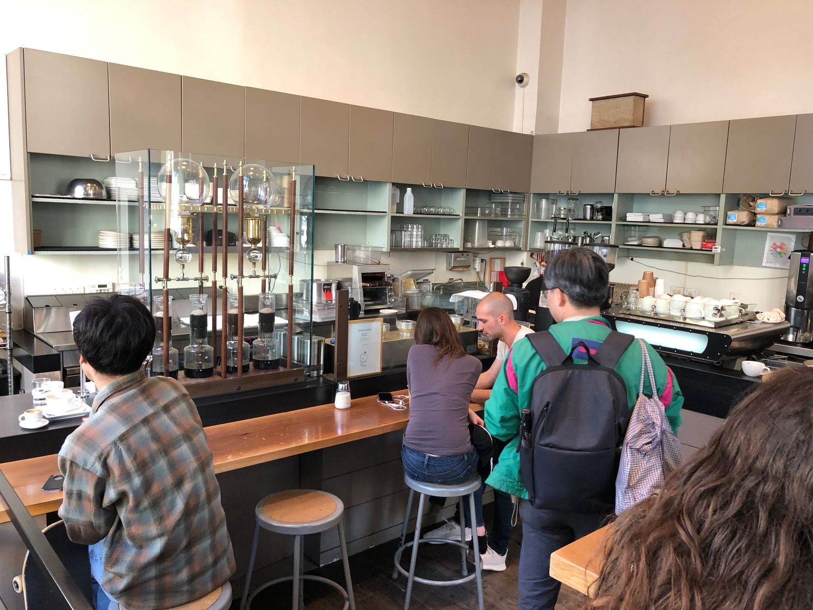 Customers at a wooden bar watching siphon coffee brewers behind glass